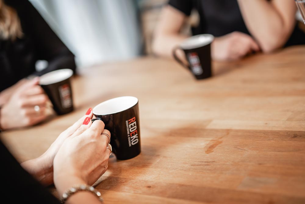 Närbild på händer från tre kvinnor som håller svarta kaffemuggar med Redekos logotyp